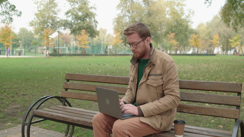 Male blogger in eyeglasses typing on laptop, sitting in park with cup of coffee