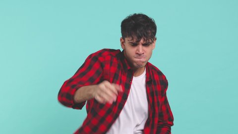 Cheerful funny smiling young man 20s wearing basic red checkered shirt posing isolated on blue turquoise color background in studio. People lifestyle concept. Dancing clenching fists waving hands