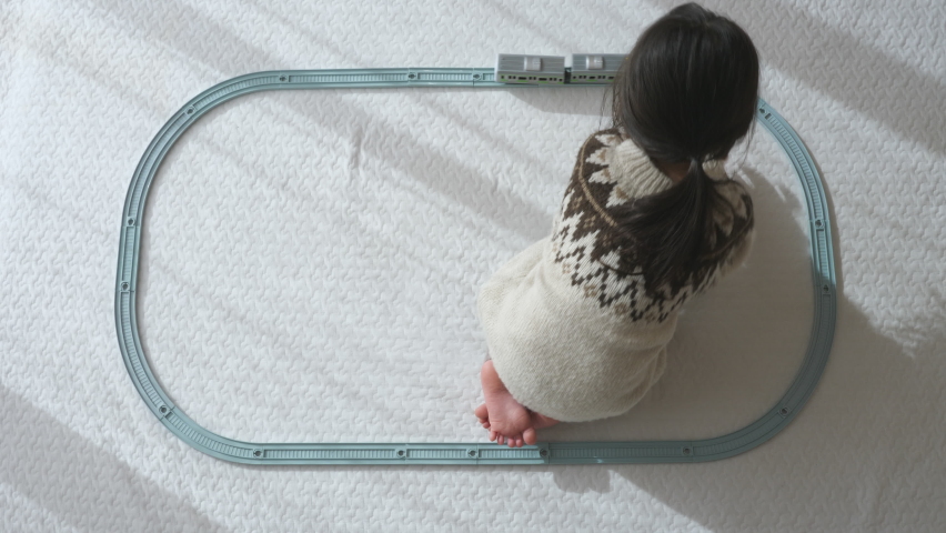Funny toddler little girl playing with a toy train connecting plastic rails on a white floor mat at home. Top view. Living room with daytime sunshine. Kid chasing a train and spinning. Cute baby child