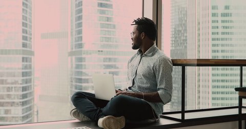 Ambitious african man sit cross-legged on windowsill in office put laptop on laps looking admires cityscape through panoramic window daydream about future, feels happy with career, position in company
