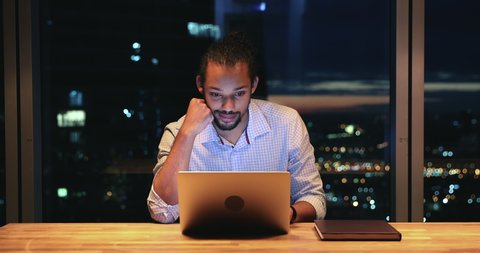 Busy 30s african ethnicity employee working hard using wireless computer stay late in modern skyscrapers office, behind his back night lit city view. Urgent project, careerism, workaholism concept