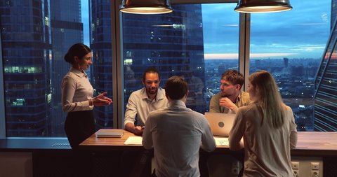 Multiethnic startup business team gathered in modern office boardroom, working until late, skyscrapers, night city view through panoramic window. Motivated ambitious workgroup brainstorming concept