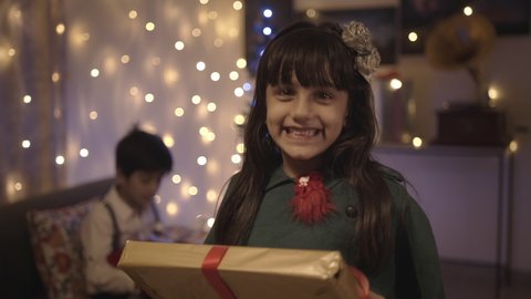 A well-dressed adorable young female kid is smiling while receiving the Christmas present in a decorated house. Happy and excited girl accepting the gift-wrapped box from parents 