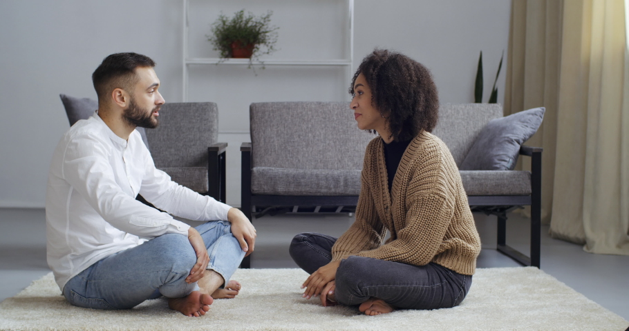 Mixed nationality couple curly African girlfriend wife and Caucasian boyfriend husband sitting together on floor on carpet in living room in new house talking dreaming planning furniture arrangement Royalty-Free Stock Footage #1063481488