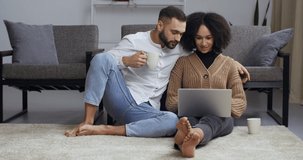 Young family multi-ethnic couple sitting embracing on living room floor looking at laptop screen reading emails watching movies video online through computer drinking caco coffee tea from white cups