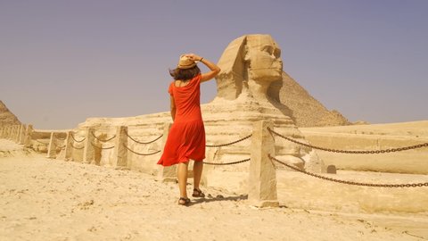 A young Caucasian brunette in a red dress and straw hat enjoying visiting the Great Sphinx of Ginza next to the pyramids of Ginza. Cairo, Egypt