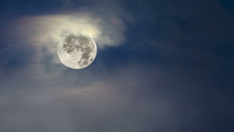 Fog moves across a full moon framed against a moody, blue sky with cloud cover.