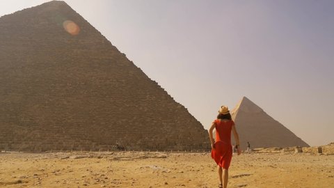 Panning shot of a young tourist in a red dress and a straw hat walking in the pyramids of Giza. Cairo, Egypt