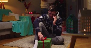 Middle plan of man sitting on floor next to sofa, calling, talking on phone next to present, while girlfriend decorating christmas tree in living room. Concept of family holidays, Christmas time.