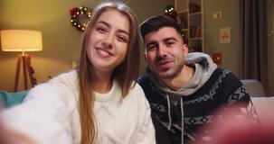 Frontal smartphone camera view of young couple husband and wife sitting on sofa, calling on webcam, video chatting, waving hand, having conversation, smiling on Christmas Eve in living room.