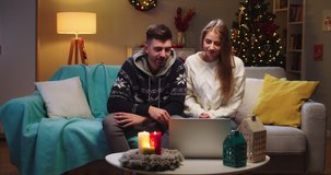 Middle plan of young couple husband and wife sitting on sofa, calling on webcam, video chatting with family using computer, waving hand, having conversation, smiling on Christmas Eve in living room.