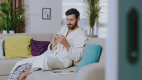 Handsome young man in domestic clothing sitting on couch smiling use phone. Feel happy. Stay at home. Social media. Communication conference portrait. Slow motion