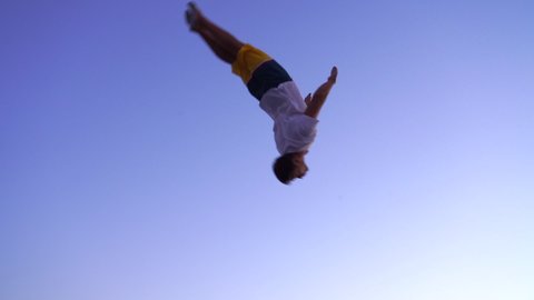Young sports acrobat jumping on a trampoline. back flip. european guy in beachwear on blue sky background