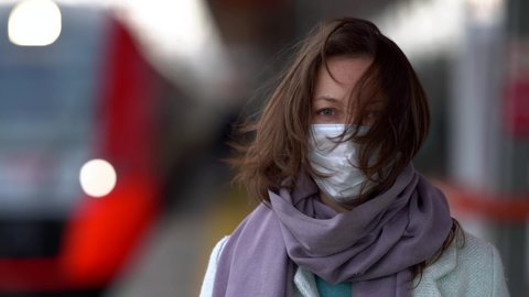 A close-up of a middle-aged woman standing on a railway platform on the street. The wind blows through her hair. She has a protective medical mask on her face. There's a train coming from behind.