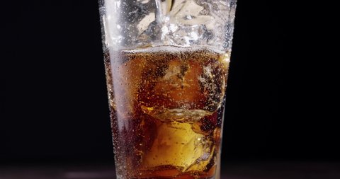 Cola is poured into a glass of cola with ice. The glass stands on a wooden table with a black background. Gas bubbles actively rise in the glass. Slow motion.