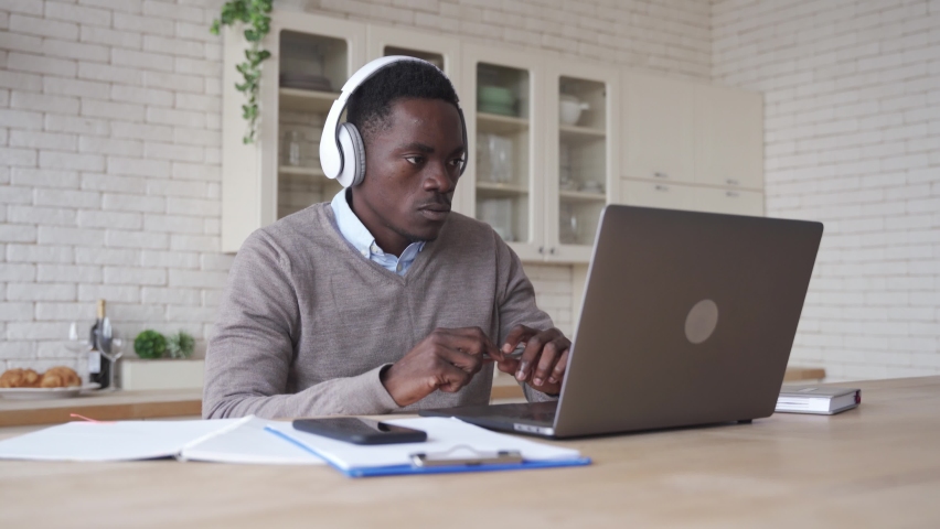 Serious african black business man, male student wears headphones talking, conference video calling, watching webinar online, social distance learning, working in web chat using laptop at home office. Royalty-Free Stock Footage #1063513525