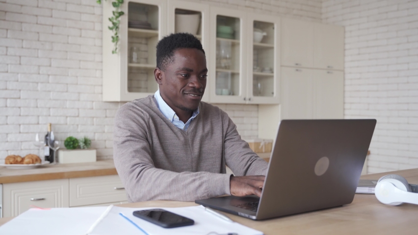 Smiling professional black african business man using laptop working from home office. Happy african male customer or student studying online, e learning, typing on computer sits at kitchen table. | Shutterstock HD Video #1063513531