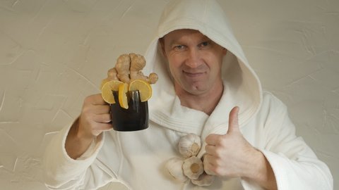 portrait. A young cheerful Caucasian man in a white coat offers to drink tea with ginger root and lemon. There is garlic on the man's neck. Virus and infection prevention concept