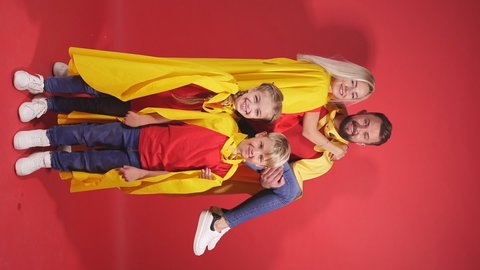 Happy parents playing superheroes with children, posing isolated over red background, wearing yellow cloaks.