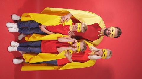 Parents and children in superhero cloaks, family of superheroes in costumes posing in studio with red background, kids and wife in the head of strong man.