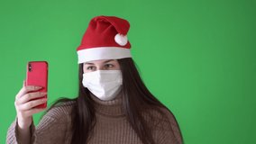 A girl in a medical mask and a Santa Claus cap talks by video using a smartphone.
