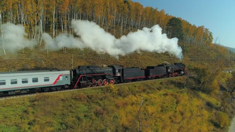 Baikal, Russia - September 16, 2019: Aerial old steam engine locomotive train rides fast on rails railroad through autumn Russia birches yellow forest. Countryside landscape. Travel tourism. Drone 4k