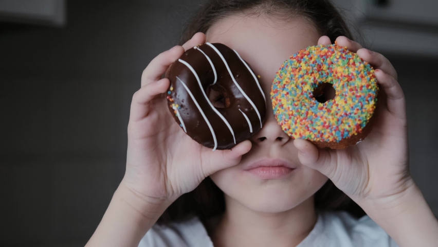 An attractive cute little girl holding bright donuts with icing in her hands, smiling, looking at the camera and bringing them to her face to look through them. Close-up