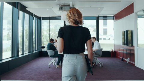 Rear view of a businesswoman walking in the board room with staff sitting around table.  Office manager arriving in the conference room with her team settling down around the table.
