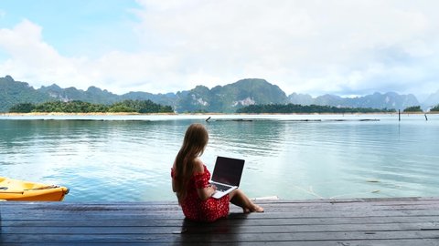 Woman Freelancer Sitting on Wooden Pier at Lake with Laptop, Relaxing and Enjoying Nature Landscape. Travel Woman with Computer in Outdoor Office on Beautiful River with Kayak and the Green Mountains.