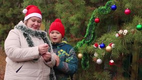 Mom and son are cheerfully shooting a clapperboard on the street. Beautiful toys on a branch. Against the background of a dense green Christmas tree. We celebrate the new year. Warm soft light.
