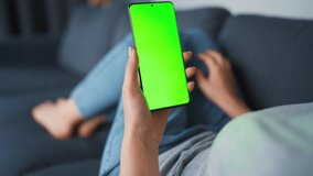 Woman at home lying on a sofa and using smartphone with green mock-up screen in vertical mode. Girl browsing Internet, watching content, videos, blogs. POV.