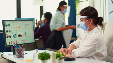 Businesspeople working wearing protection face masks in office room during coronavirus. Team in new normal business financial company typing on pc, checking reports, analysing data looking at desktop