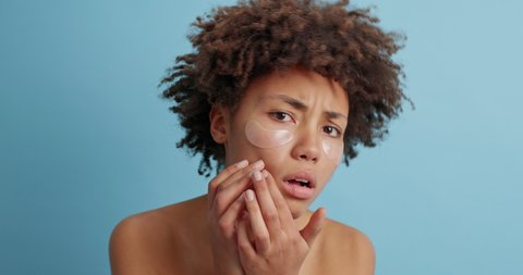 Stressed surprised woman notices how many pimples she has squeezes acnes on face looks in terror at camera presses on skin problem zone wears beauty patches poses indoor naked wants perfection