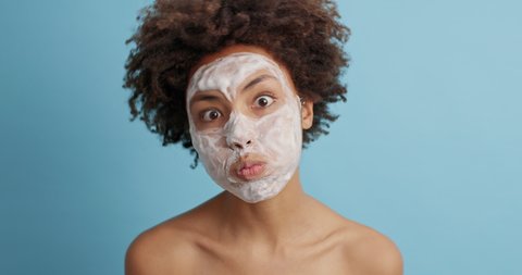 Curly young woman makes funny grimace pouts cheeks has fun wears beauty face mask for healthy smooth skin undergoes cosmetic facial treatment stands bare shoulders isolated on blue background