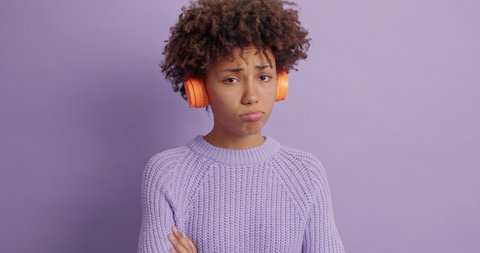 Disappointed dark skinned young woman listens audio track tries to improve her spoiled mood looks unhappily at camera wears sweater isolated over purple background. Negative emotions concept