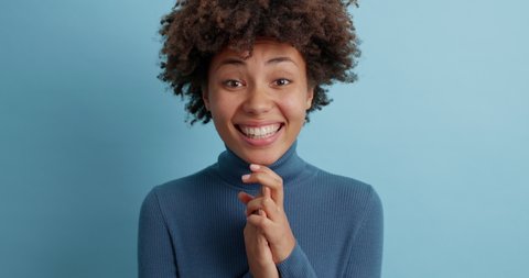 Cheerful glad Afro American woman wears casual turtleneck does winner gesture clasps hands and smiles broadly celebrates success wears casual clothes isolated over blue background. Triumph concept