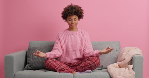 Calm relaxed dark skinned woman meditates with hands spread sits in lotus pose practices yoga breathes deeply stays at home meditates on comfortable sofa wears casual clothes isolated on pink wall