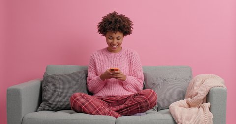 People technology and leisure concept. Relaxed curly haired young Afro American woman uses smartphone types text messages dressed in casual clothes sits on comfortable sofa with cushions around