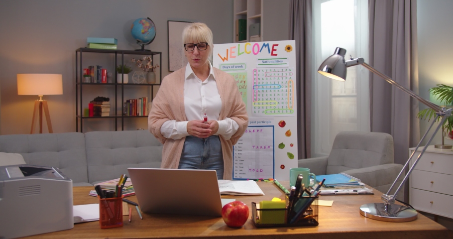 Confident Caucasian woman in glases having online class on laptop at home. Adult female English teacher writing days of week on white board and speaking on camera. Education, profession.