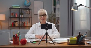 Successful adult woman in glasses sitting at table and teaching students via video conference indoors. Caucasian female professor using smartphone to talk and writing in notebook. Education.