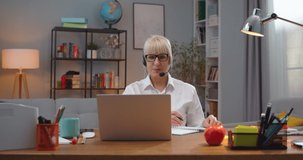 Beautiful Caucasian woman in glasses sitting at table and examining students via video conference chat. Adult female school teacher in headphones taking notea and talking at home. Education concept.