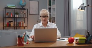 Charming Caucasian woman sitting at table and working indoors. Tired adult female teacher shutting laptop computer screen and putting off glasses. Job, education, technology concept.