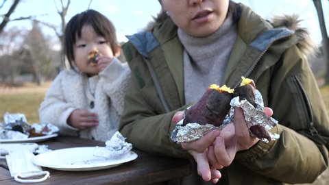 Asian mother halves delicious freshly baked sweet potatoes with both hands outside. Background of a little toddler girl watching it while eating sweet potatoes. Autumn winter outdoors. Happy family