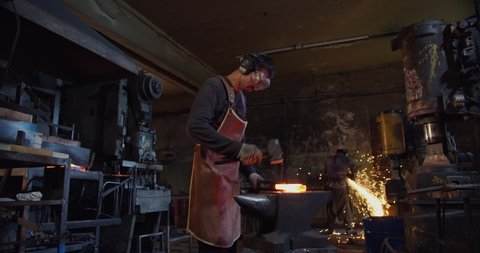 In a general shot, in an atmospheric forge, a blacksmith in a leather apron and goggles takes the red-hot metal from the furnace with metal tongs, puts it on the anvil, and hits it with a hammer