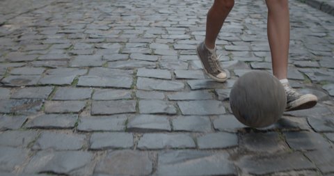 Crop view of teenager feet dribbling at empty old city street with paving stones. Person in sneakers running and kicking football ball. Concept of lifestyle, childhood, sport.