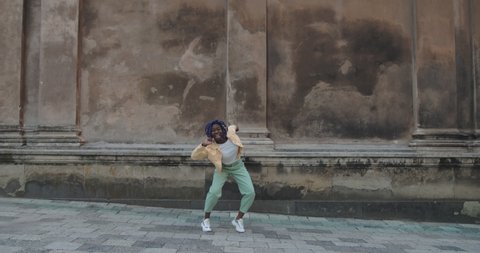 Attractive millennial female person dancing while walking along old city street. Smiling young woman with dreadlocks looking happy while moving in rhytm. Concept of emotions. Outdoors.