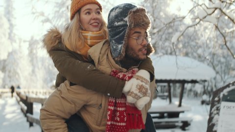 Young cheerful man giving piggyback ride to joyous girlfriend while walking through snowy park on winter day