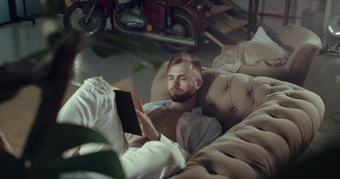 a man lying on the couch reads a book and relaxes at home. A poor man learns and flips through a book with useful information. Slow motion.