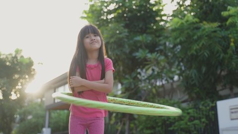 Young Asian little girl playing hulahoop exercise outdoor at home. Young cute kid wear cusaul clothes, enjoy swing the hula hoop with happiness and smile enjoy working out on street for health care.