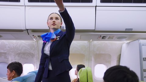 Beautiful Caucasian flight attendant walking on aislel to close luggage compartment and check the readiness of airplane before take off. Safety check procedure of cabin crew in the plane.
